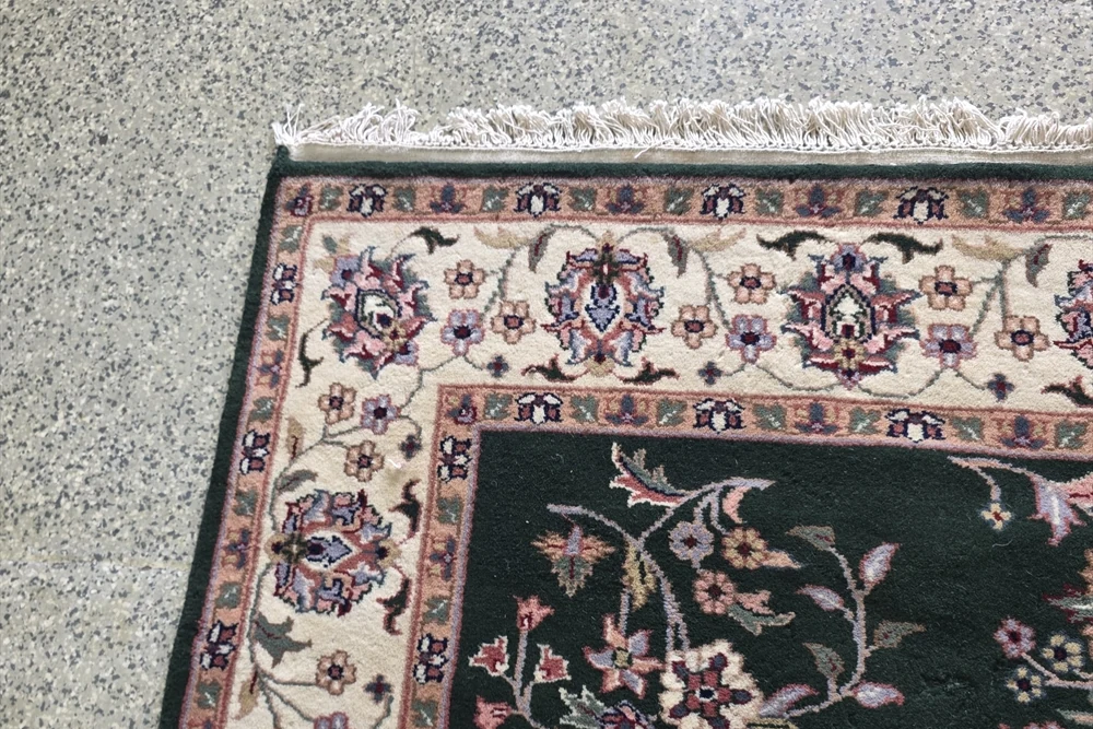 Corner of a dark-green floral rug showing white fringe, the end finish and the bound side edge.