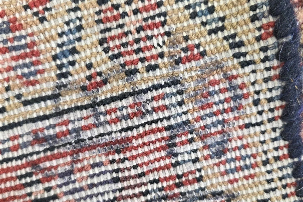 Close-up of a rug’s reverse showing rows of cream, red, gold and dark yarn beside a dark bound edge.